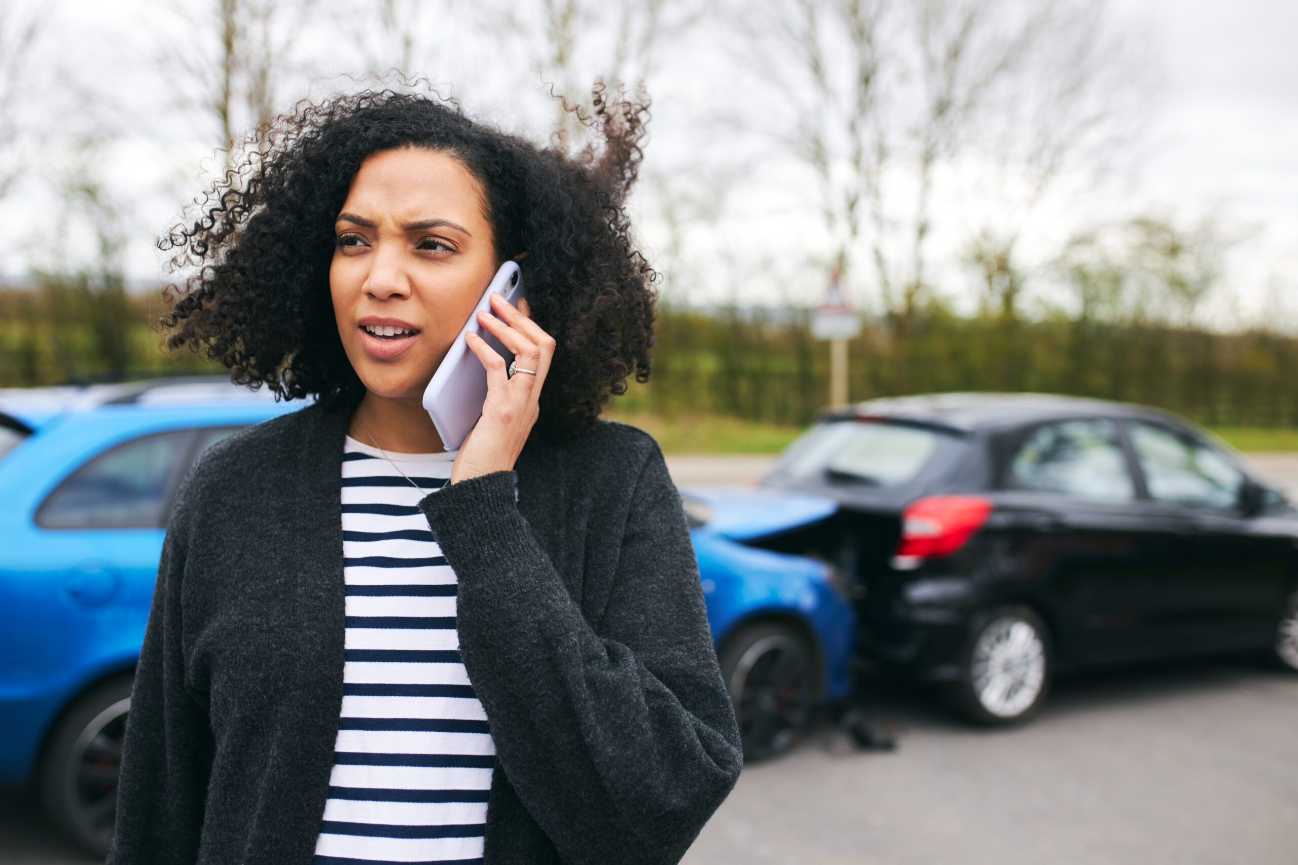 Young woman standing by damaged car after traffic accident reporting incident to insurance company using mobile phone - – write off claims assistance in the UK